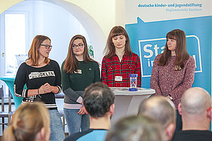 vier Mädchen halten eine Präsentattion vor einer Gruppe Erwachsener