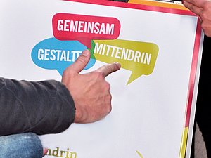 Eine Hand zeigt auf das Logo von Gemeinsam Mittendrin Gestalten auf einem Plakat.