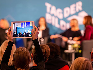 Ein Tablet wird in die Luft gehalten. Auf dem bildschirm sieht man die Bühne mit dem Tag der Bildung Logo.